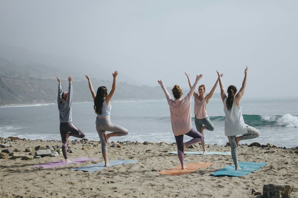Un viaje en grupo de WeRoad con cinco mujeres haciendo la postura del árbol sobre esterillas de yoga en una playa de arena frente al océano.