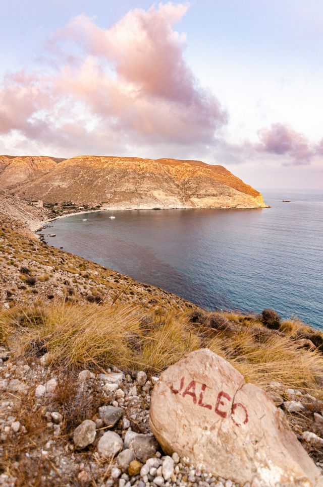 Una roca pintada con la palabra 'JALEO' se asienta en una colina seca y cubierta de hierba, con vistas a una cala con veleros y acantilados áridos al atardecer.