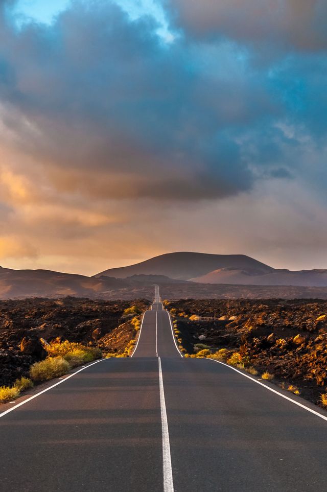 Una strada diritta si estende attraverso un paesaggio vulcanico roccioso verso le montagne sotto un cielo nuvoloso al tramonto.