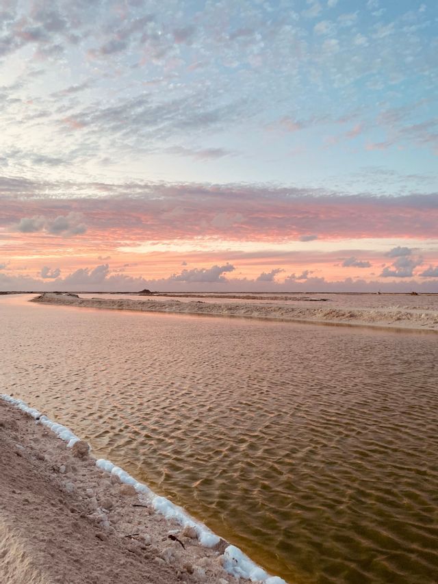 Salt pans with channels of water reflecting a colorful sunset sky with pink and blue clouds.