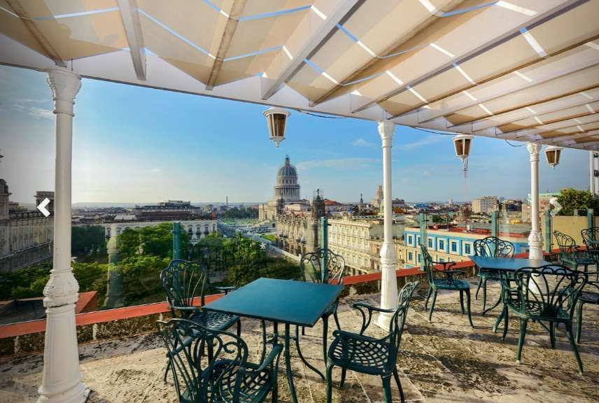 Una terrazza panoramica con tavoli e sedie in metallo verde si affaccia su un paesaggio urbano con un grande edificio a cupola sotto un cielo blu.