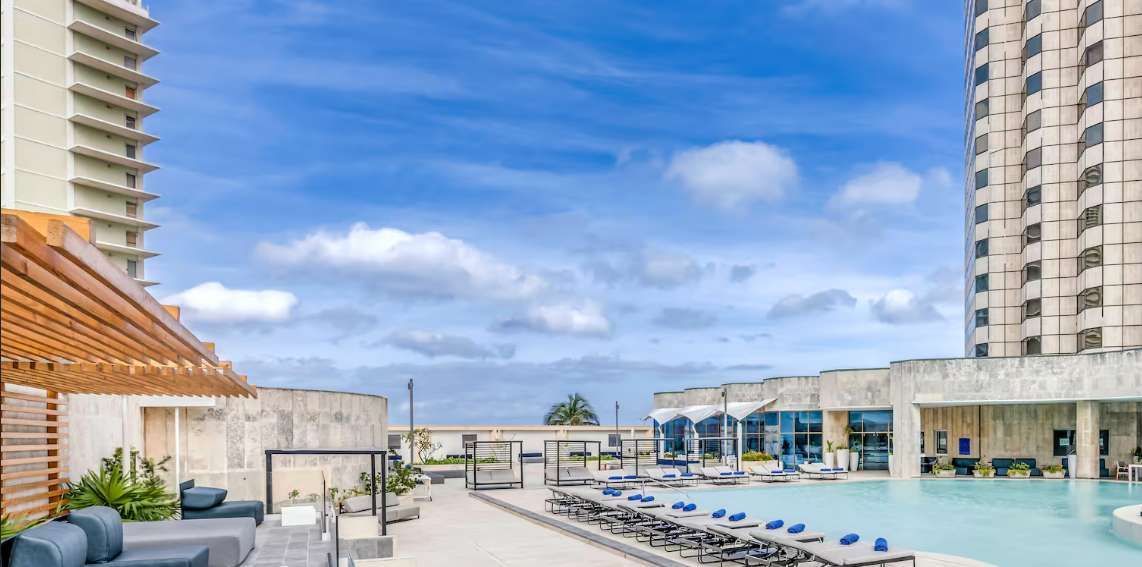 Un'area piscina panoramica sul tetto con lettini vuoti e moderni edifici alberghieri, sotto un cielo parzialmente nuvoloso.