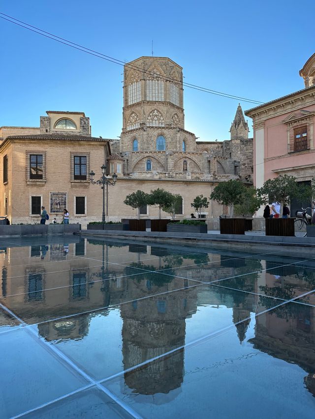 Un campanile storico in pietra si riflette sulla superficie di una fontana bassa e a griglia in una piazza.