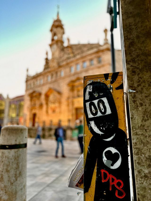 Un adesivo di street art con un personaggio a fumetti nero e un cuore è su un palo, con un edificio storico sfocato sullo sfondo.