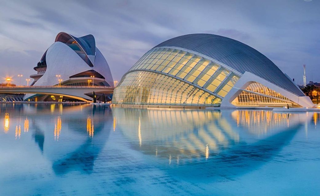 Due moderni edifici architettonici illuminati al crepuscolo, le cui luci si riflettono sull'acqua blu calma in primo piano.