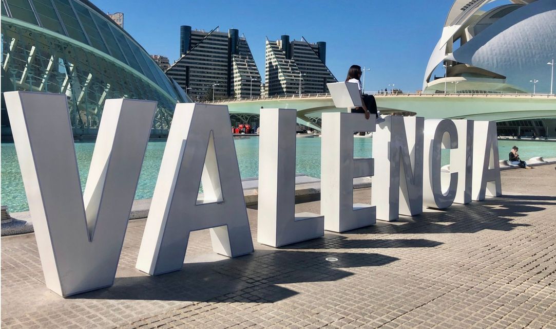 Grandi lettere bianche che formano VALENCIA su una piazza pavimentata, con una donna seduta sulla lettera 'E' sotto un cielo limpido e soleggiato.