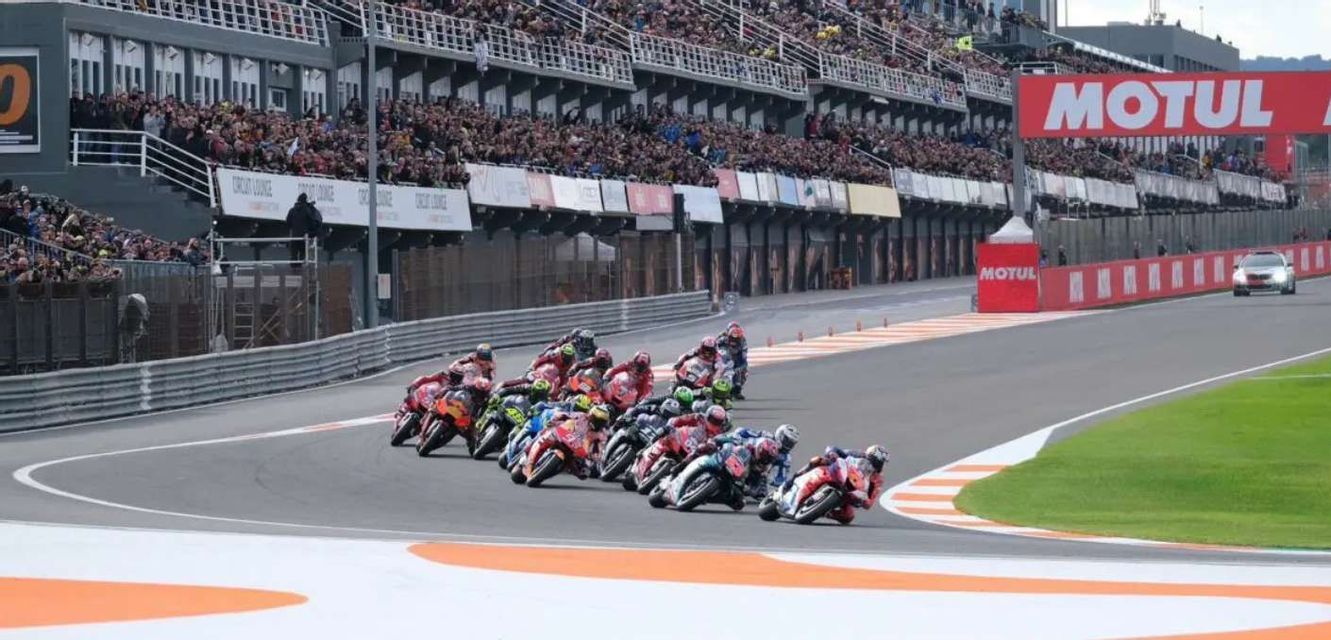 Un gruppo di motociclisti si inclina in una curva stretta su una pista, con una grande folla che guarda dalle tribune.