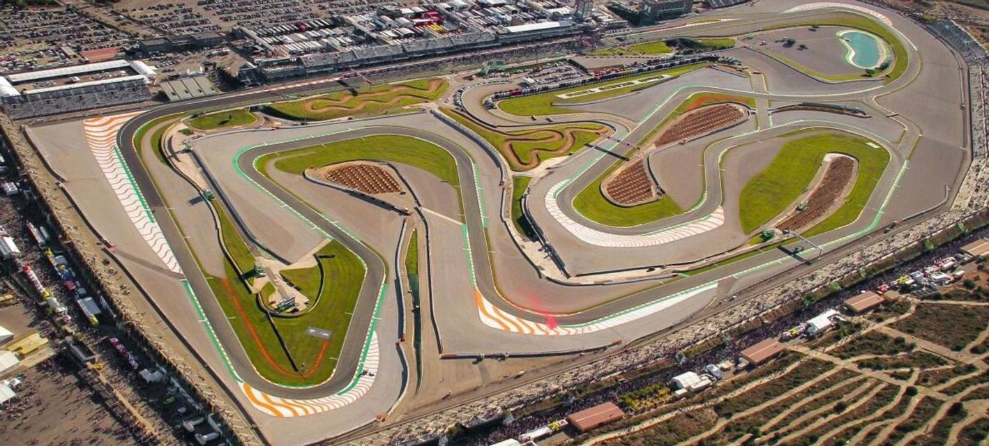 Vista aerea di un ampio e sinuoso circuito automobilistico con curve multiple, tribune e aree verdi interne.