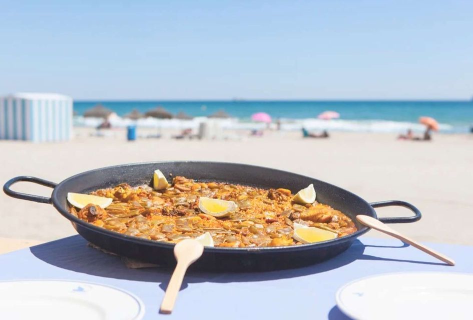 Una padella di paella con spicchi di limone su un tavolo in una spiaggia assolata, con l'oceano sullo sfondo.