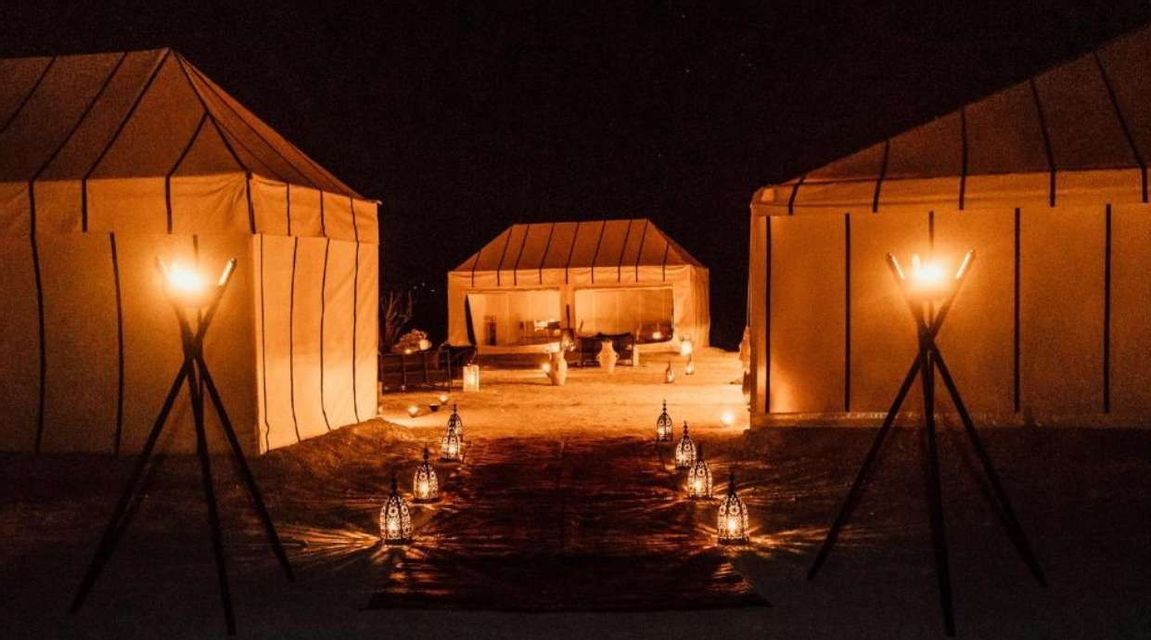 Faroles y antorchas iluminan un camino entre grandes tiendas de lona en un campamento en el desierto durante la noche.