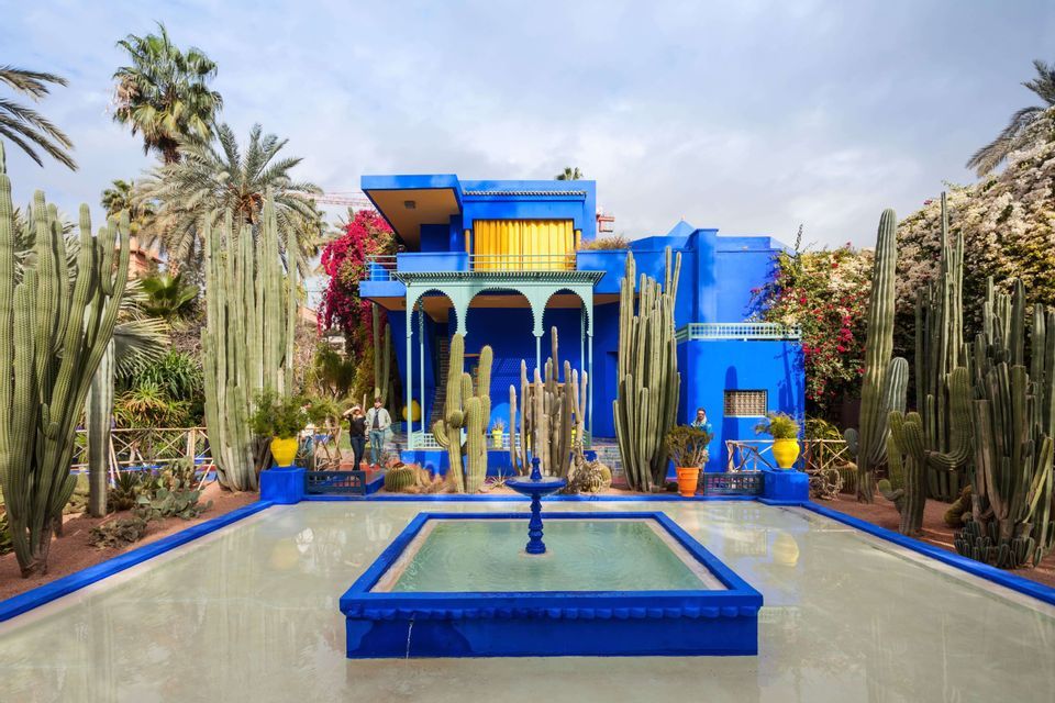 Un bâtiment bleu éclatant se dresse au cœur d'un jardin de grands cactus et de palmiers, avec un bassin d'eau et une fontaine à l'avant.