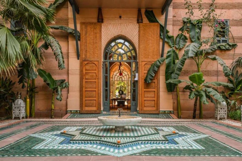 A star-shaped fountain with green and white mosaic tiles sits in a Moroccan-style courtyard with an arched doorway and large potted plants.