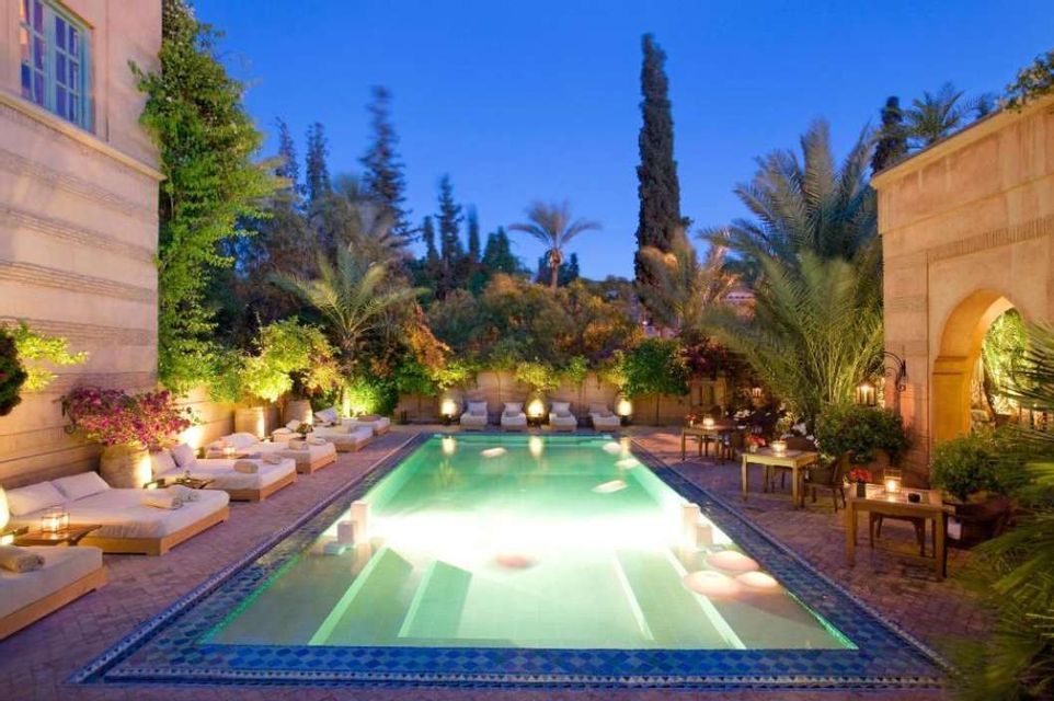An illuminated rectangular swimming pool in a courtyard at dusk, surrounded by lounge chairs and lush plants.