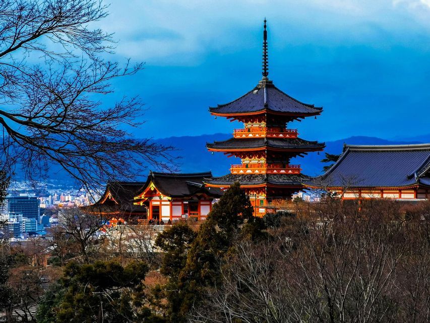 Una pagoda e un tempio giapponese arancioni a più livelli, su una collina che domina una città, incorniciati da alberi spogli sotto un cielo azzurro.