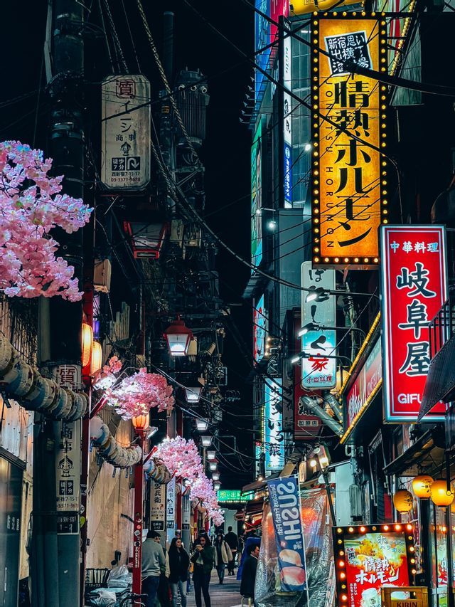 Una stretta strada cittadina di notte, illuminata da luminose insegne al neon giapponesi, con persone che camminano sotto e fiori di ciliegio rosa in alto.