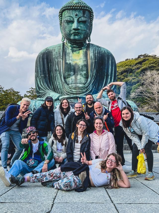 Un gruppo WeRoad in viaggio posa e sorride per una foto davanti a una gigantesca statua bronzea del Buddha seduto sotto un cielo parzialmente nuvoloso.