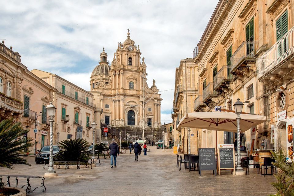 La gente attraversa un'ampia piazza pavimentata verso una cattedrale barocca ornata con una grande cupola.