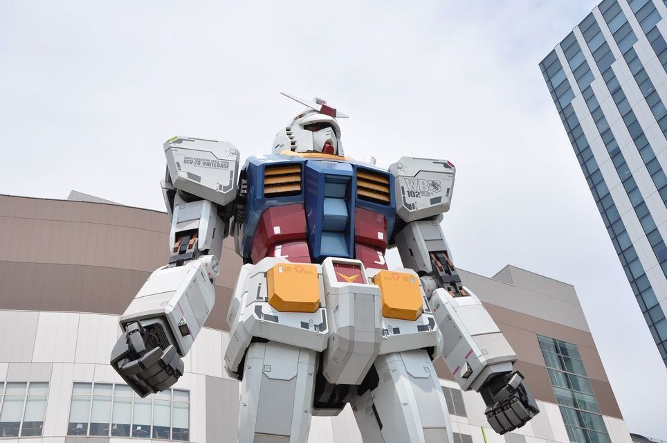 Una inquadratura dal basso di una statua gigante a grandezza naturale del robot Gundam, posta di fronte ad alti edifici moderni.