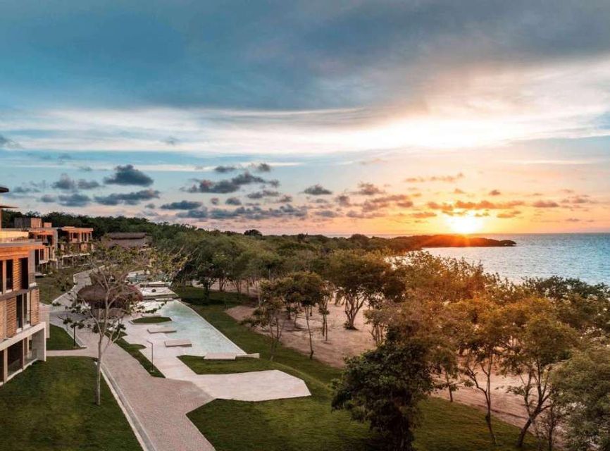 An aerial view of a coastal resort with modern buildings, pools, and trees overlooking the ocean at sunset.