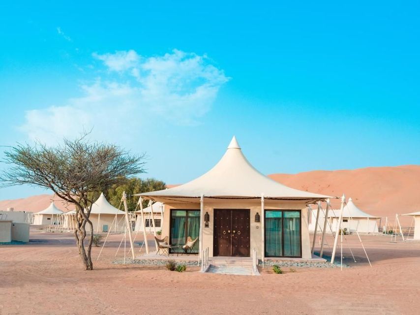 Una tenda glamping di lusso con tetto a punta si trova in un campo nel deserto, con dune di sabbia sullo sfondo sotto un cielo blu.