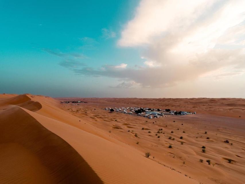 Un insediamento di edifici bianchi è adagiato tra le dune di sabbia arancione in un vasto deserto, sotto un cielo turchese con nuvole.