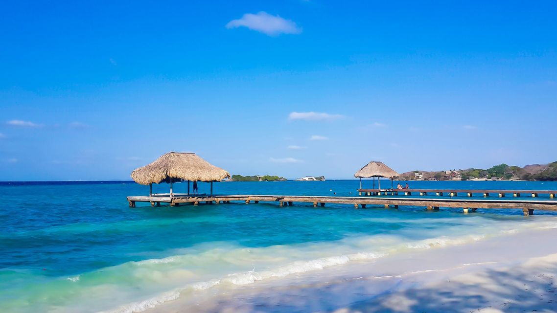 A wooden pier with two thatched-roof huts extends over turquoise ocean water from a white sand beach under a clear blue sky.