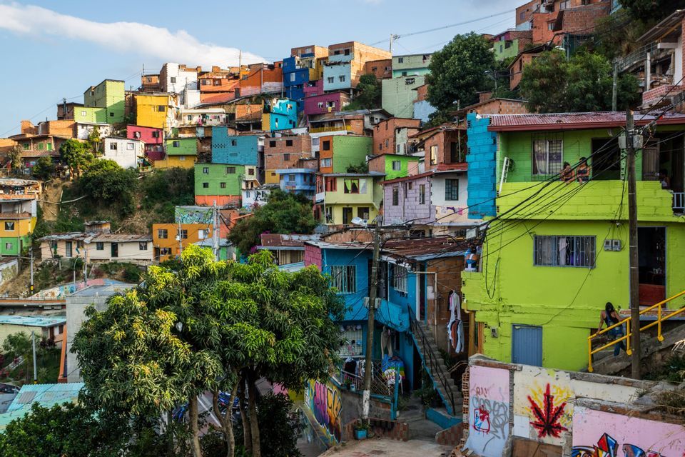 A dense neighborhood of brightly colored houses built on a lush green hillside under a partly cloudy sky.