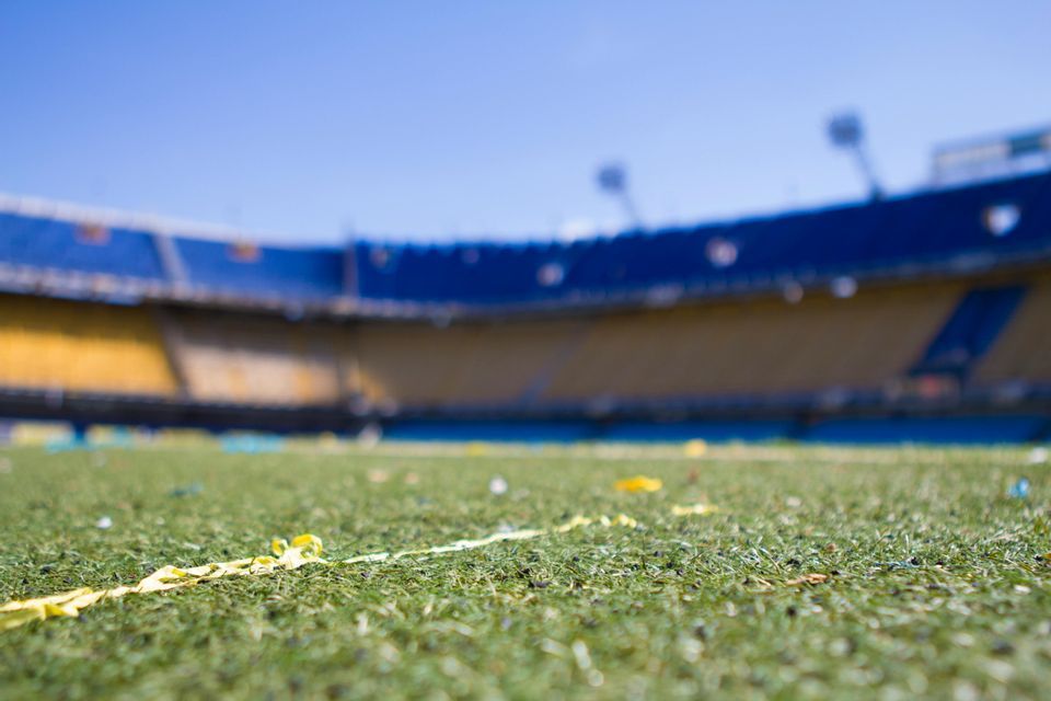 Primer plano en contrapicado de confeti amarillo sobre el césped de un estadio, con gradas azules y amarillas borrosas al fondo.