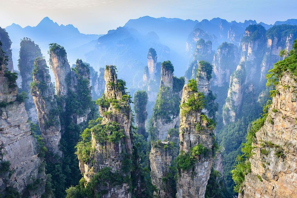 Alte formazioni rocciose a pilastro coperte di vegetazione verde si ergono da una valle nebbiosa con montagne sullo sfondo.