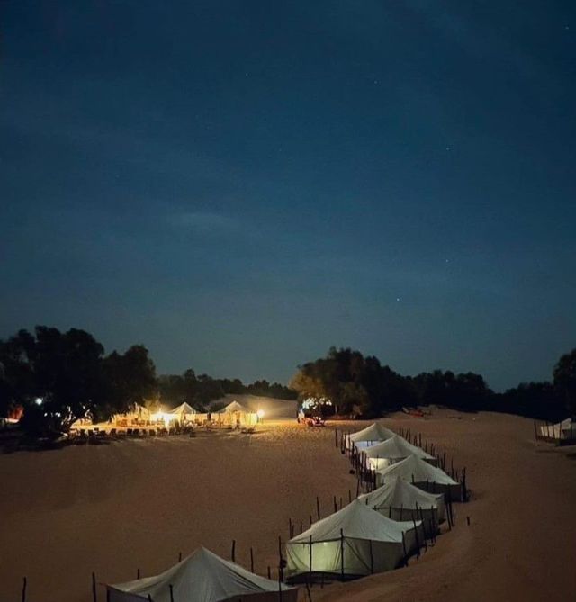 Tiendas blancas iluminadas forman un campamento en dunas de arena del desierto bajo un cielo nocturno estrellado.