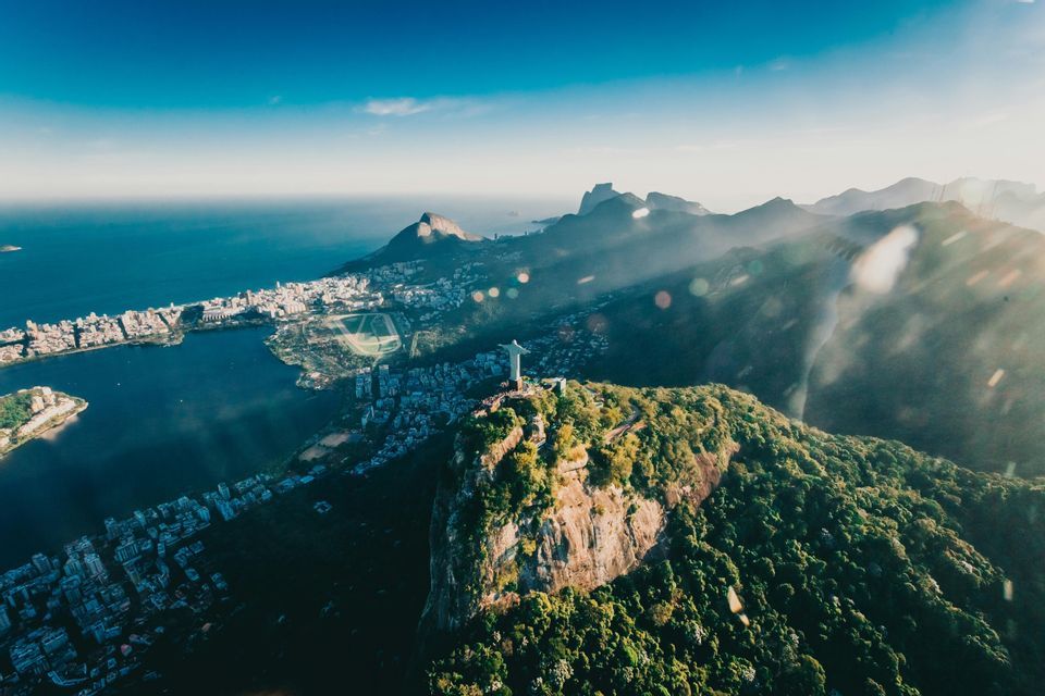 Una vista aerea della statua del Cristo Redentore su una montagna, che domina una città e la costa sotto un cielo azzurro.