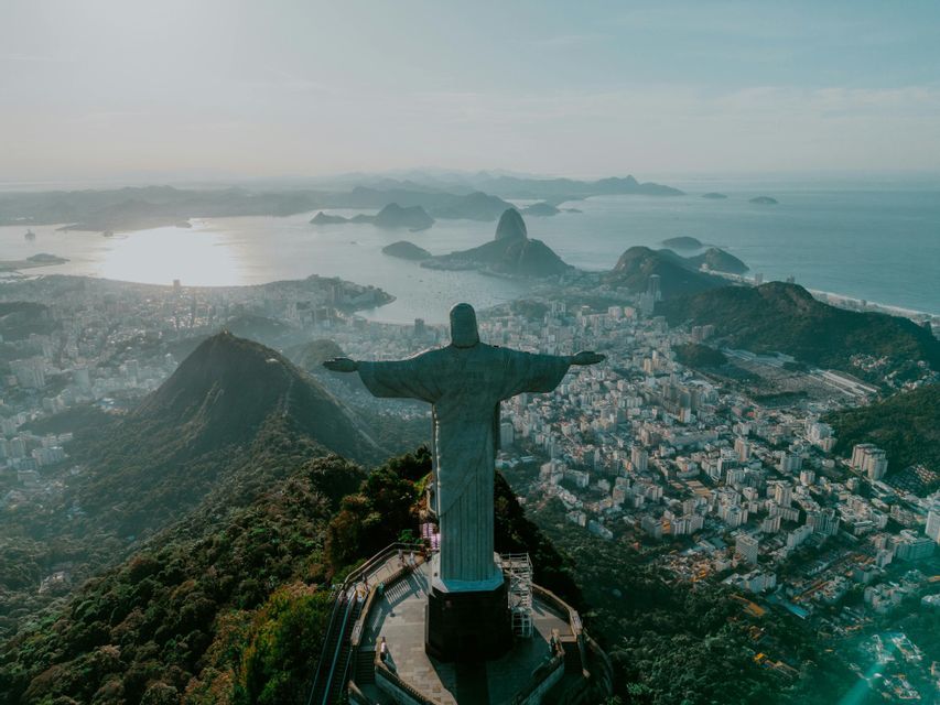 Una vista aerea da dietro una grande statua con le braccia distese, che si affaccia su una vasta città costiera, baia e montagne all'alba.