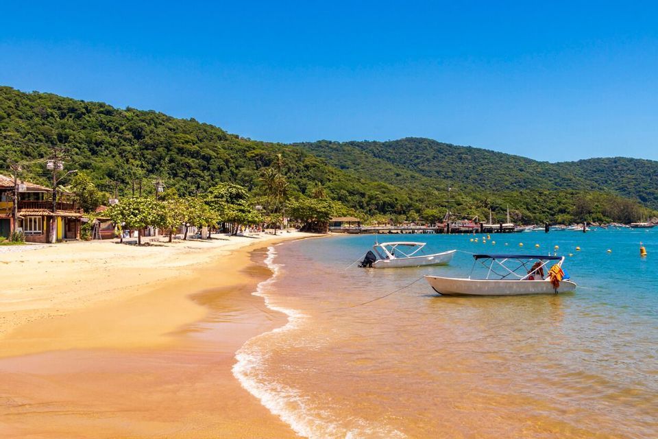 Due piccole barche sono ancorate nelle acque limpide di una baia tranquilla, accanto a una spiaggia sabbiosa alberata e con alle spalle una grande collina boscosa.