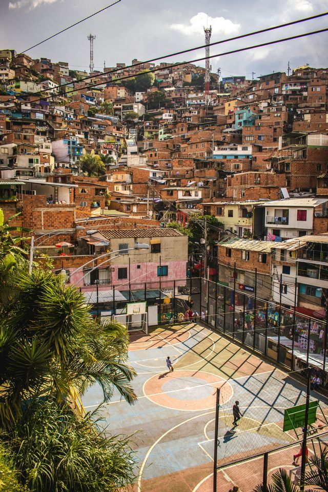 Vista dall'alto di un campo da basket recintato con persone che giocano, situato in una fitta comunità collinare di case in mattoni.