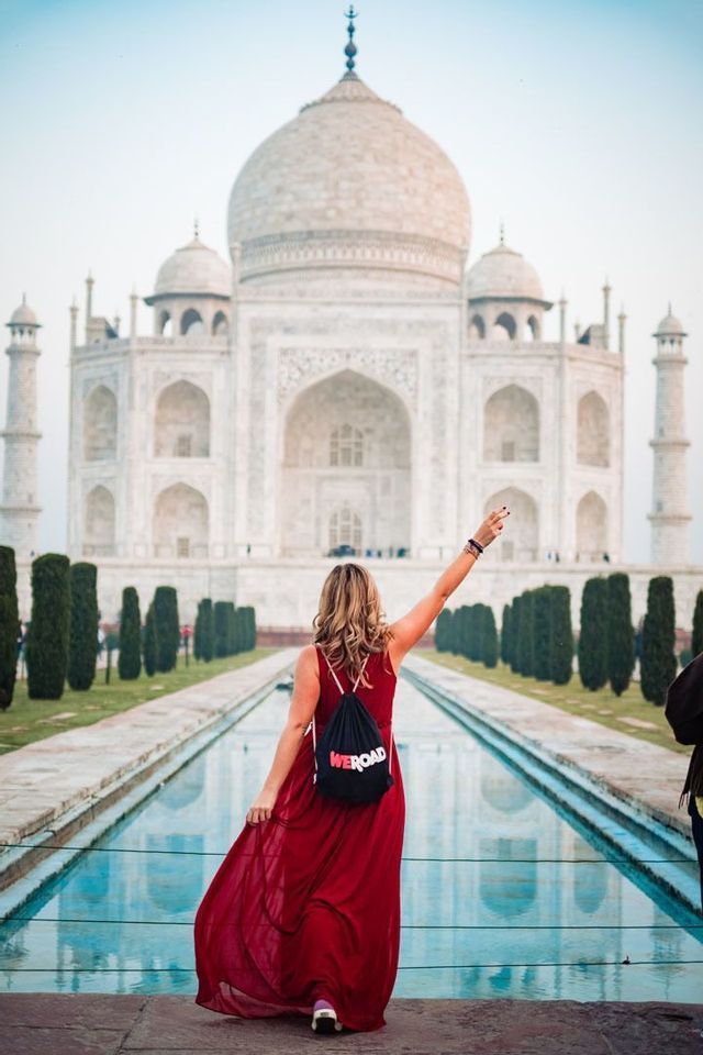 Una mujer de espaldas con un vestido rojo largo y una mochila WeRoad, levanta el brazo frente al Taj Mahal y su estanque.