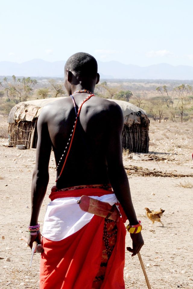 Un homme en tenue traditionnelle rouge, vu de dos, tient un bâton et contemple un paysage aride avec une cabane.