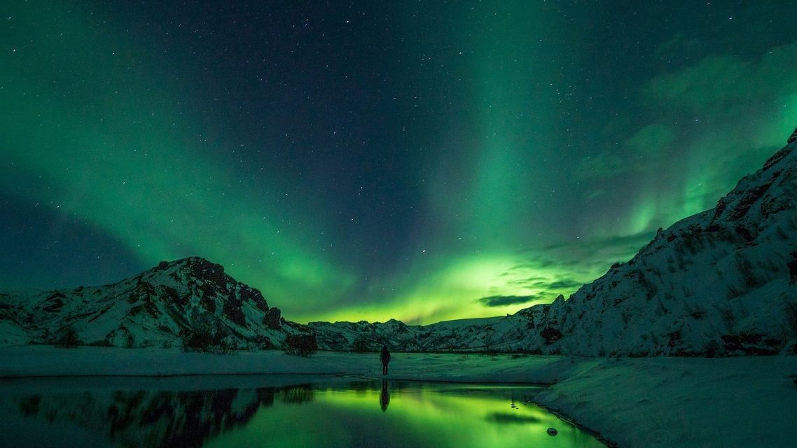 Eine Person beobachtet die Nordlichter über schneebedeckten Bergen, deren grünes Leuchten sich im ruhigen Wasser eines Sees spiegelt.
