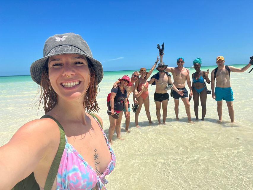Eine lächelnde Frau mit Fischerhut macht ein Selfie mit einer WeRoad-Gruppenreise im seichten, klaren türkisfarbenen Wasser stehend.