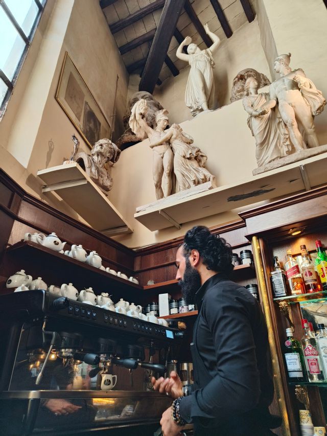Un barista barbudo prepara café en una máquina de espresso en un café con paredes adornadas con estatuas clásicas y teteras.