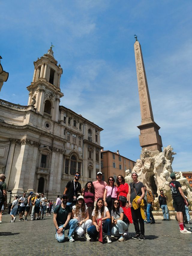 Un viaje en grupo de WeRoad posando para una foto en una plaza histórica y soleada con un obelisco y una fuente de fondo.