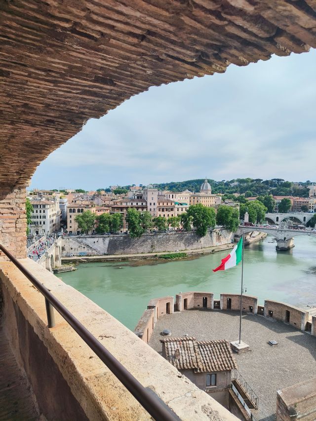 Una bandera italiana ondea en el torreón de una fortaleza que domina una ciudad europea con un río ancho y puentes, vista desde debajo de un arco de ladrillo.