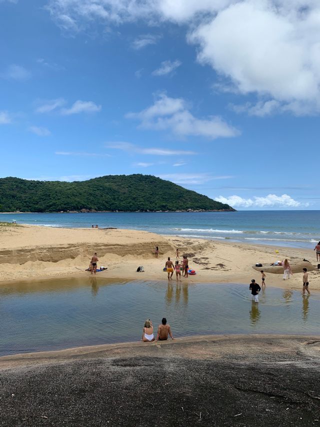 Menschen entspannen an einem Sandstrand mit einem Gezeitenbecken, einem grünen Hügel und dem Meer im Hintergrund.
