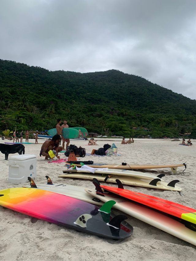 Tavole da surf colorate giacciono su una spiaggia sabbiosa con un gruppo WeRoad che prepara la propria attrezzatura, con una grande collina verde sullo sfondo.
