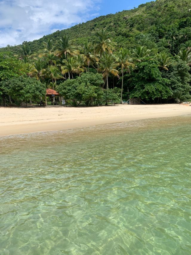 Ein Blick über klares, flaches Wasser zu einem Sandstrand mit dichtem Tropenwald und einem üppig grünen Hügel unter blauem Himmel.