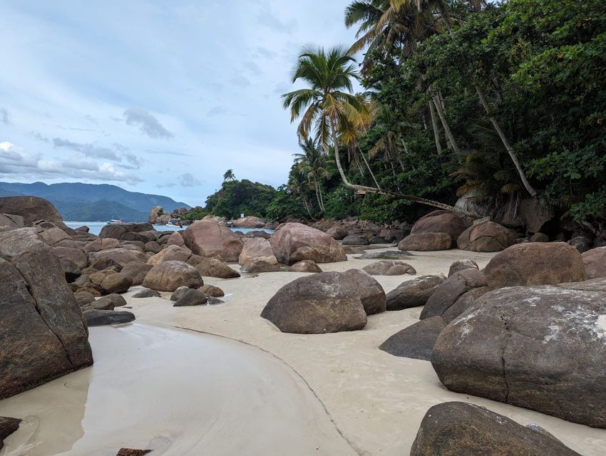 Große, abgerundete Felsbrocken liegen an einem Sandstrand neben einem tropischen Wald mit Palmen, mit fernen Bergen auf der anderen Seite des Wassers.