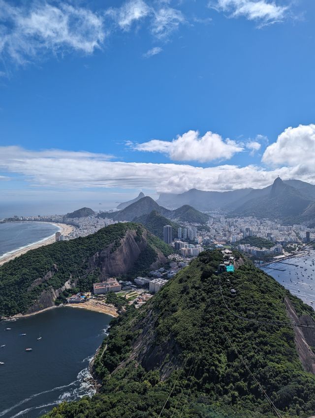 Vue plongeante d'une ville côtière avec des plages et des montagnes verdoyantes bordant l'océan sous un ciel bleu et nuageux.