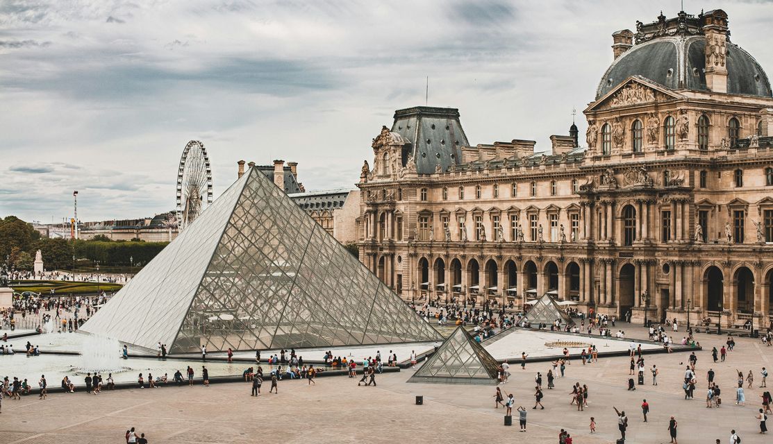 Crowds of people gather in a grand courtyard around a large glass pyramid, with a historic building in the background.