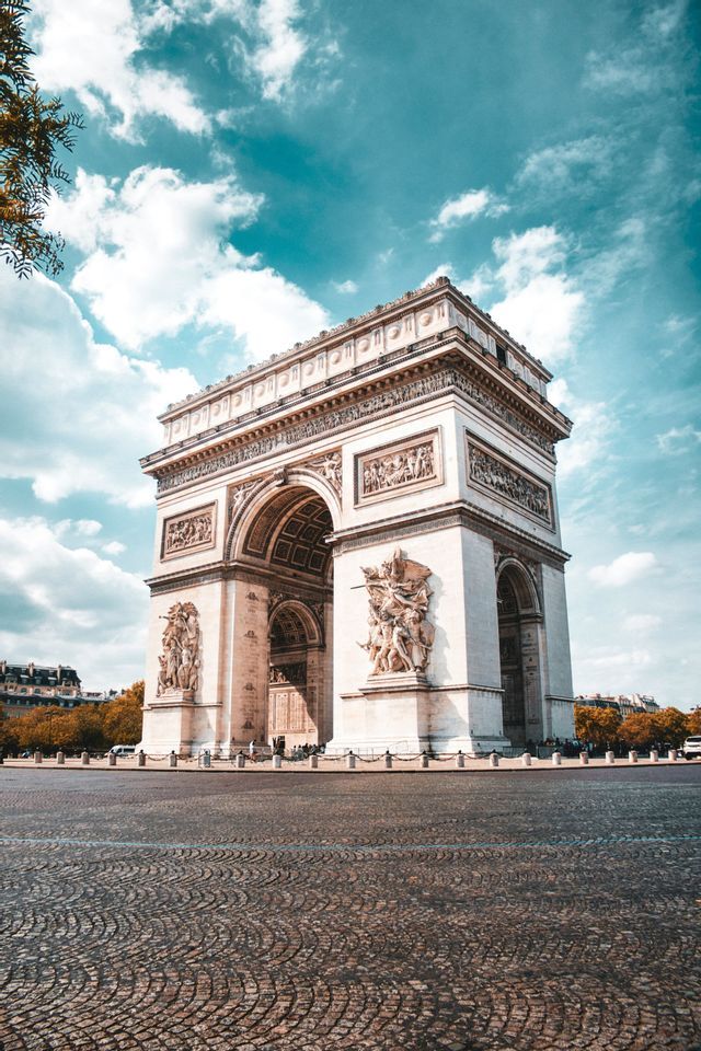 Un grande e ornato arco trionfale si erge su un'ampia piazza lastricata sotto un cielo azzurro con nuvole sparse.