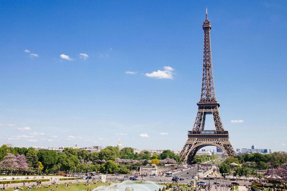 A daytime view of the Eiffel Tower from a park, with people relaxing on the grass and a city skyline behind it.