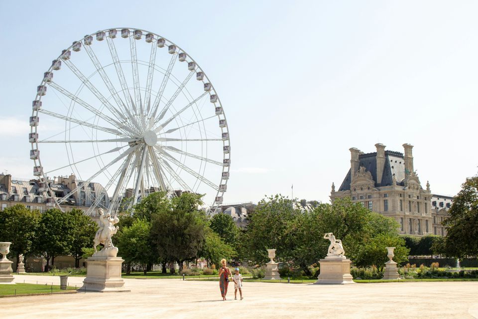 Ein Erwachsener und ein Kind gehen unter klarem Himmel durch einen großen Park mit Statuen, einem großen Riesenrad und historischen Gebäuden.
